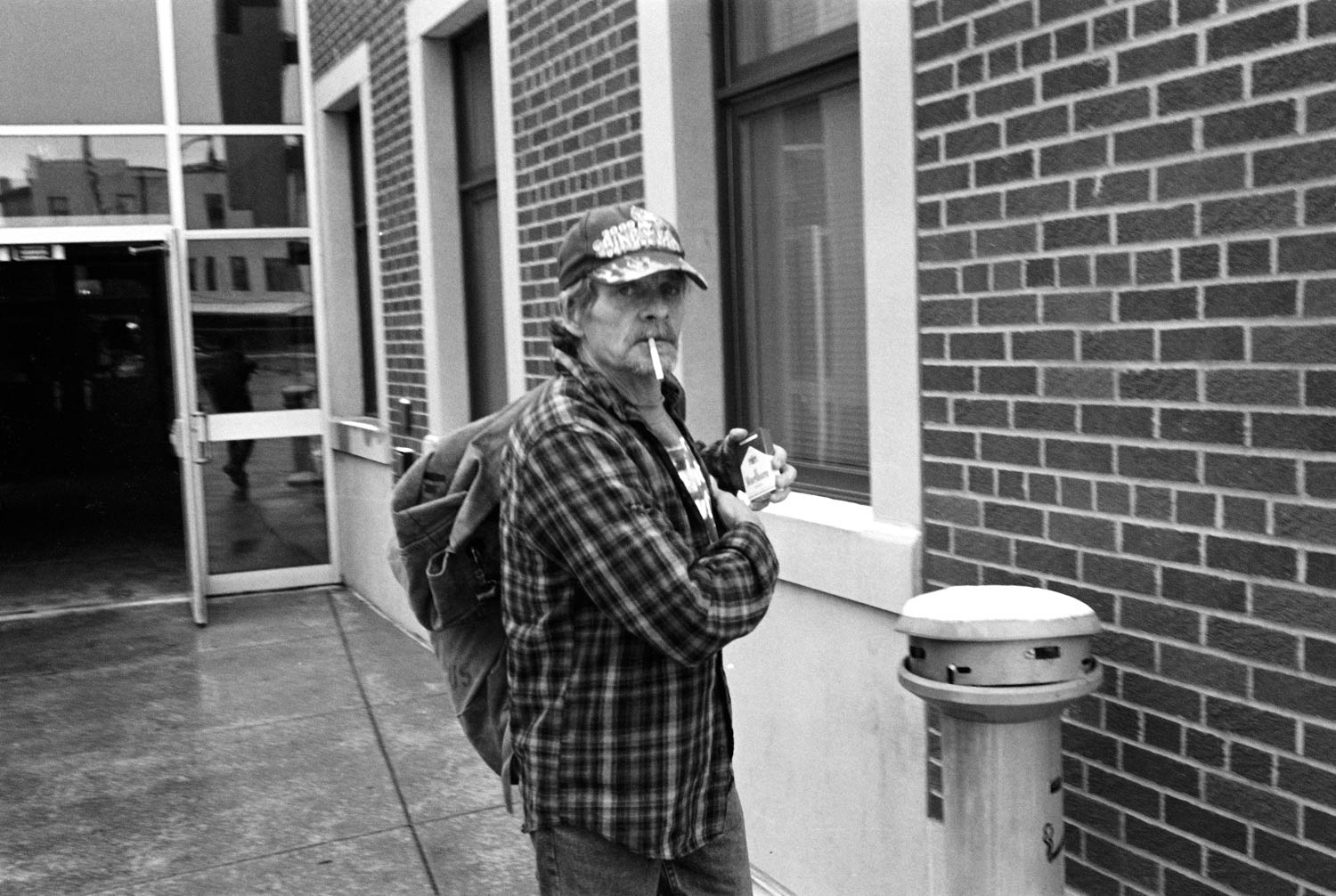 A black and white picture of a man putting a carton of cigarettes back into a flannel shirt pocket while walking out of the B.C. Junction in Binghamton, NY.