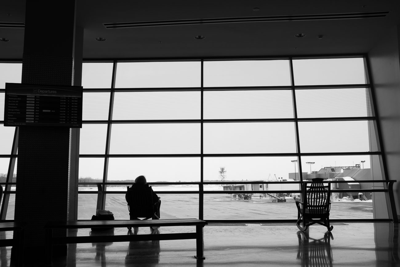 Black and white photo of a man sitting in a rocking chair in front of the large windows of the Syracuse International Airport.